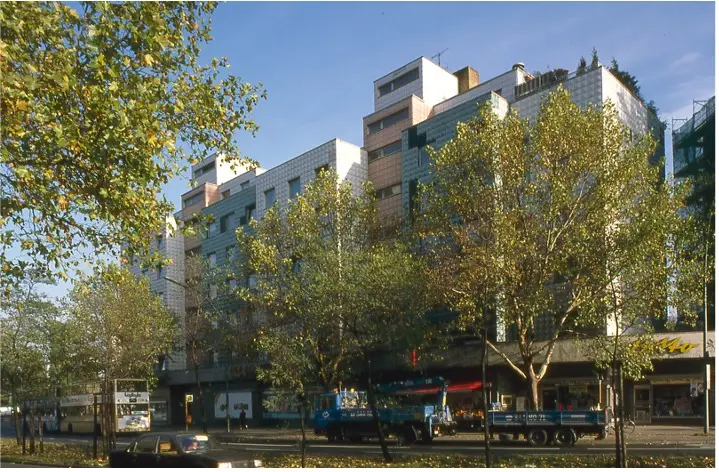 Eine Straßenszene in der Stadt mit einer Reihe mittelhoher Gebäude, jedes mit einer modernen Fassade. Bäume säumen den Bürgersteig, ihre grünen Blätter verdecken teilweise die Gebäude. Ein blauer Lastwagen parkt auf der Straße und der Himmel ist klar und blau.